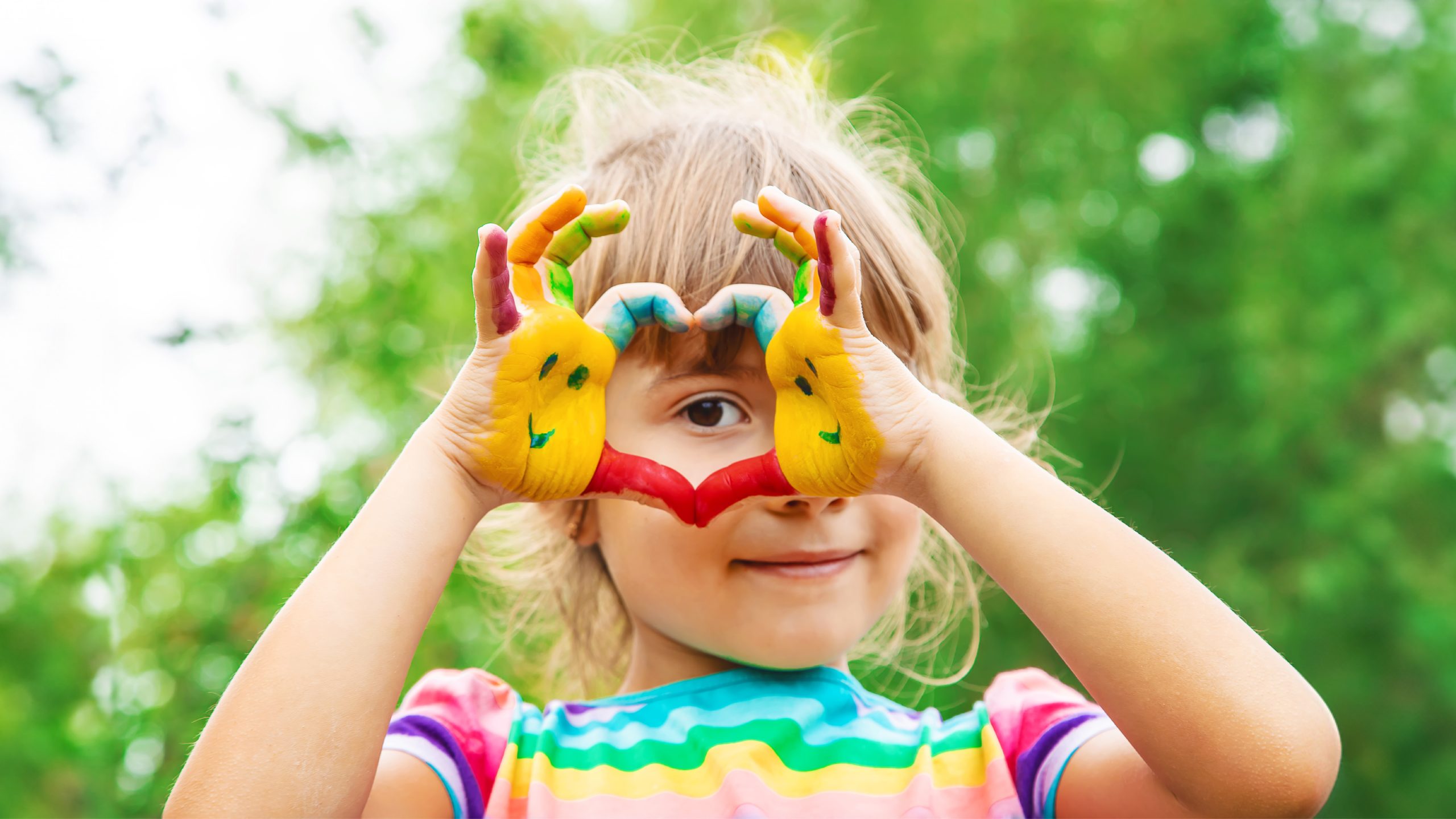 Photograph of a smiling child with painted hands forming a heart shape, representing creativity and self-expression.